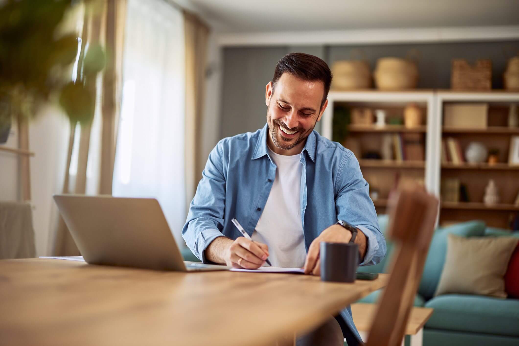 Man happily writing notes at home with laptop and coffee.